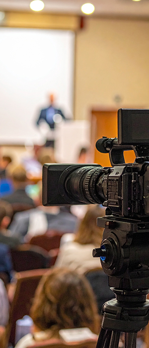 Aufzeichnung eines Vortrags. Im Vordergrund eine Kamera, der Mittel- und Hintergrund ist unscharf. Im Publikum Menschen von hinten, vor einer Bühne sitzemd. Im Hintergrund ein Mann vor einem Rednerpult, dem Publikum zugewandt, dahinter eine weiße Leinwand.