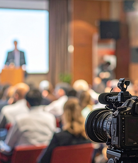 Aufzeichnung eines Vortrags. Im Vordergrund eine Kamera, der Mittel- und Hintergrund ist unscharf. Im Publikum Menschen von hinten, vor einer Bühne sitzemd. Im Hintergrund ein Mann vor einem Rednerpult, dem Publikum zugewandt, dahinter eine weiße Leinwand.